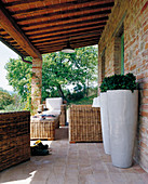 Veranda of Tuscan country house with terracotta tiles, comfortable wicker furniture and two white floor vases in foreground