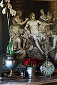 Flowers in ornamented metal vases on shelf below picture with Ancient Greek motif