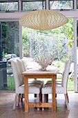 Chairs with white upholstery at wooden table and pale, basketwork pendant lamp in front of terrace doors with view of garden