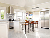 Spacious, white kitchen with long kitchen counter, stainless steel door fronts and high wicker stools at counter