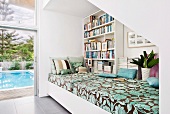 Daybed with patterned cover against bookcase on wall and next to terrace door with view of pool