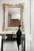 Schwarze Katze springt von Wandtisch aus verspiegeltem Glas, an der Wand Spiegel mit geschnitzten Blumenmotiven auf hellem Holzrahmen