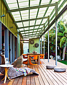 A glass-roofed veranda with comfortable wicker loungers and a large wooden dining table with orange plastic chairs