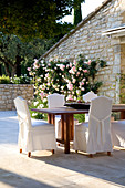 Hussenstühle und Holztisch auf Terrasse mit rankendem Rosenstrauch an der Natursteinfassade eines französischen Landhauses