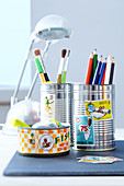 Coloured pencils and paintbrushes in decorated tin cans on a desk