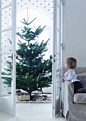 Blick auf Weihnachtsbaum durch offen stehende Flügeltür und Kind auf Sofa