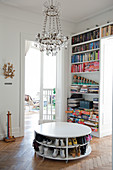 Shoes stored on shelves of white, round, low table on herringbone parquet floor and chandelier in front of bookcase next to open door leading to living room