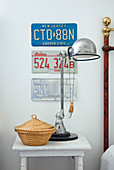 Retro table lamp and wicker basket with lid on vintage bedside table below American car number plates on wall