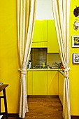 Yellow-painted foyer with gathered curtains at open doorway with view into modern fitted kitchen