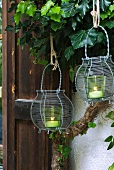 Two green tealight holders in wire baskets hanging in front of ivy-covered house facade