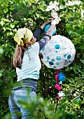 Woman hanging rice paper lantern decorated with round stickers and pompoms in garden