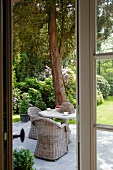 View through terrace door of wicker chairs and stone table with flowering garden in background