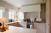 Simple, country-house kitchen with pale wooden doors and porthole window next to cooking area with built-in extractor hood