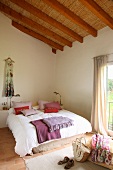 Exposed roof structure with straw mats in pleasant, bright bedroom; red and purple cushions and blankets on French bed