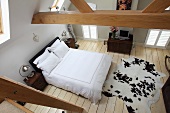 View from steep gallery staircase of wooden roof beams: birds' eye view of double bed with snow-white covers and cow-skin rug on wooden floor