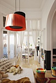 Soft toys on sofa, colourful stools and red and black pendant lamps in child's bedroom with open glass door leading to playroom in background