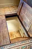 Halboffene Kellerluke in Fussboden mit gemusterten Fliesen und Blick auf steile Treppe