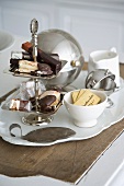 Biscuits and packaged chocolate bars on a tiered cake stand next to assorted sugar bowls on a vintage porcelain platter