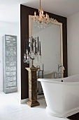 Free-standing vintage bathtub and multi-armed candelabra on antique plinth in front of floor-to-ceiling mirror on partition wall and modern, metal chest of drawers