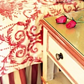 Artificial roses on bedside table painted white and red next to bed with traditional patterned bedspread