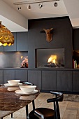 Elegant designer dining area in front of dark wall of fitted cupboards with integrated fireplace below deer-head sculpture