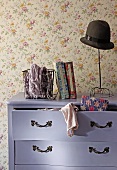 Old hat on vintage hatstand on top of Baroque chest of drawers painted purple with clothing spilling from open drawer against wall with floral wallpaper