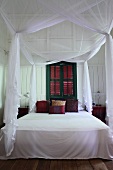 Airy, white fabric over canopied bed in front of green-painted window with closed shutters