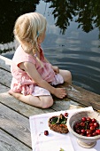 Little blond girl with cherries by the side of a pond