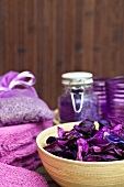 Wellness und Entspannung - violette Blüten in Bambusschale, Badesalz und Lavendelsäckchen im Hintergrund