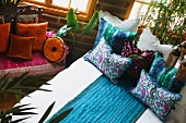 Kissen mit folkloristischem Stoffmuster auf Bett neben Holzbank mit Kissen in Orangetönen