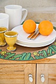 Tablett mit Orangen auf weißem Tellerstapel neben Eierbechern und Trinkbechern auf halbhohem Holzschrank