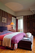 Doppelbett mit Bettwäsche in verschiedenen Rottönen und Sitzbank am Bettende in elegantem Schlafzimmer im Landhausstil