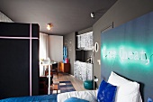 Bett an Wand mit montierter Leuchtschrift in offenem Schlafbereich und Blick auf Kochnische neben Essplatz