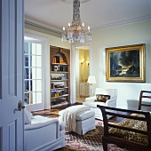 Wohnzimmer mit Lüster, großem goldgerahmtem Landschaftsbild und einem eingebauten Wandregal; im Vordergrund ein Stuhl im Empirestil