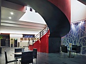 Lobby with small tables and groups of four chairs below impressive, curved staircase with red flank wall