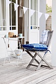 Teak chair with blue and white cushion and bistro table on greyed wooden terrace; open glass door and bunting hung diagonally in background