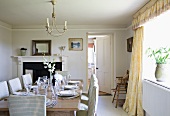 Elegant, country-house dining room in pale, Nordic shades with festively set wooden table, chairs with checked upholstery and curtains with pelmet; open fireplace and open door leading to kitchen in background.