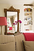 Mirror with antique-style gilt frame behind beige sofa; lampshade and folded blanket in a rich pink; feminine atmosphere in living area