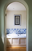 View through open arched doorway of wood-clad bathtub with tiled splashback in shades of pale blue and framed picture on wall
