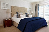 Small wooden chest of drawers and table lamp in English maritime style next to double bed with blue and white bed linen and tall, upholstered headboard