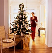 Decorated Christmas tree on trunk next to dish of biscuits on antique armchair in doorway and view of woman in grand living room
