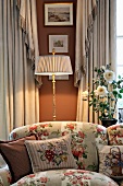 Cushions on rose-patterned sofa in front of illuminated, antique-style standard lamp between elegant curtains and against russet-painted wall with framed landscapes