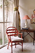 Rustic, red wooden chair next to glass wall and small writing desk with traditional table lamp in corner of room
