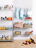 Crockery and drinks on metal kitchen shelving next to baskets and kitchen utensils hanging on perforated mounting panel