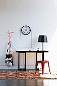 Red metal stool on rug in front of console table, black table lamp and wall clock