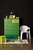 Green stool in front of green-painted chest of drawers next to white plastic chair against black wall