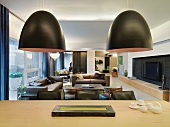 Designer hanging lamps with black shades above a dining table made of light wood in front of an elegant living room suite