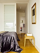 Bed with shiny bedspread and simple wooden stool in front of open sliding door with view of bathroom