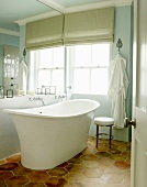 View through open door of free-standing vintage bathtub on honey-comb patterned floor tiles in front of window with Roman blind