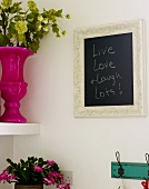 Framed blackboard hanging on a wall next to a shelf with a potted plant and flower vase